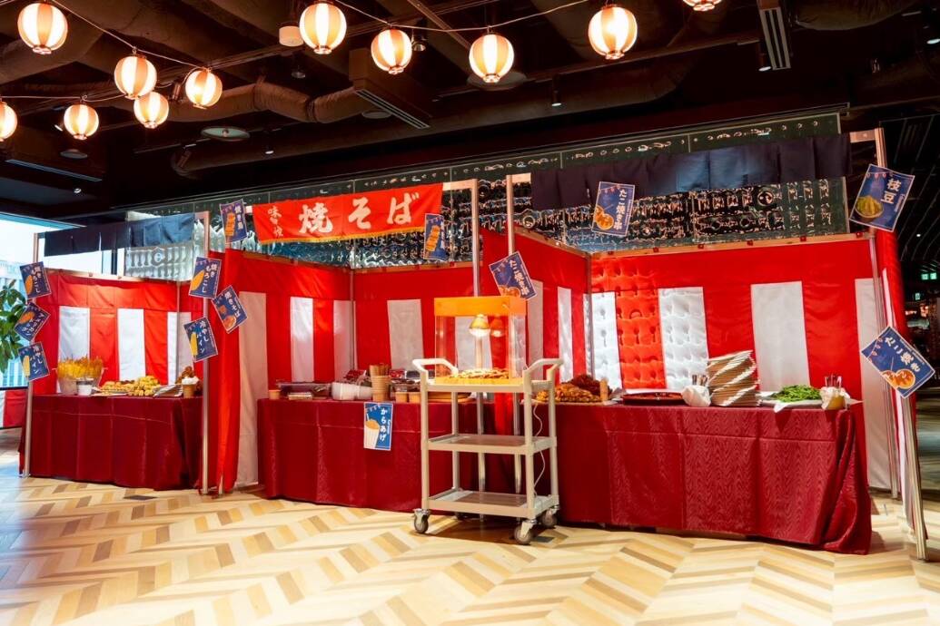 Festival food stalls with red-and-white striped curtains, tables laden with dishes, and a metal serving cart in the center under hanging lanterns.